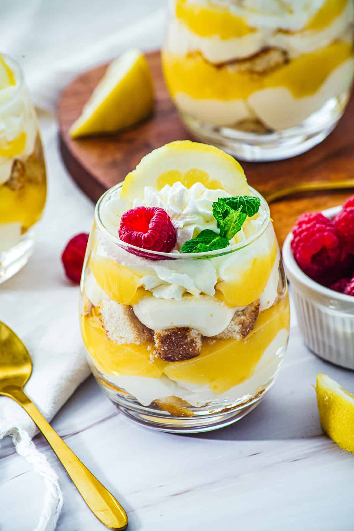 A glass dessert with lemon curd, cream, and cake, topped with raspberry, mint, and whipped cream—these delightful Lemon Cheesecake Trifle Cups are the perfect blend of tangy and sweet in every spoonful.