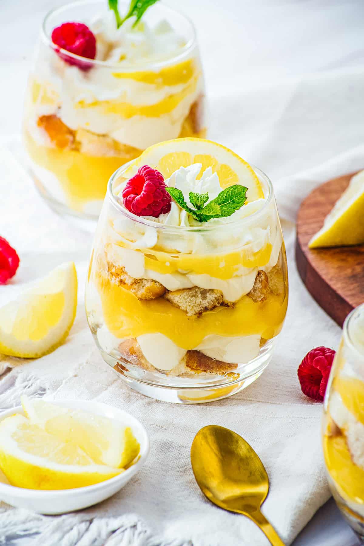 Layered Lemon Cheesecake Trifle Cups in a glass, topped with whipped cream, raspberry, mint, and a lemon slice.