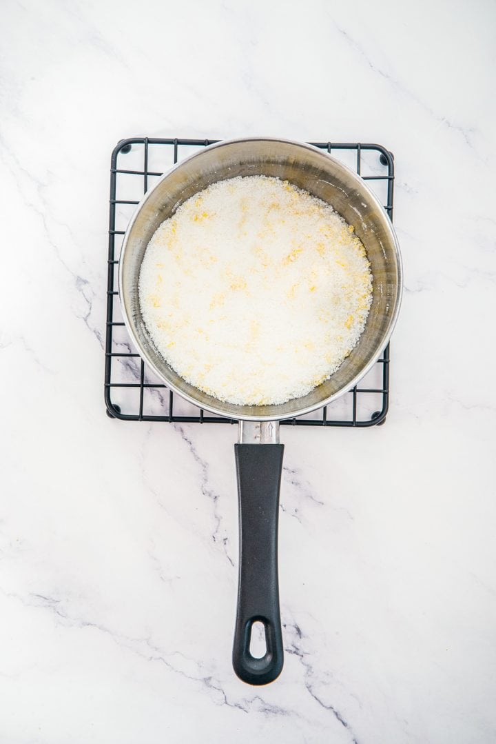 A saucepan with granulated sugar sits on a wire rack over a white marble surface, ready for the next step in preparing Lemon Cheesecake Trifle Cups.