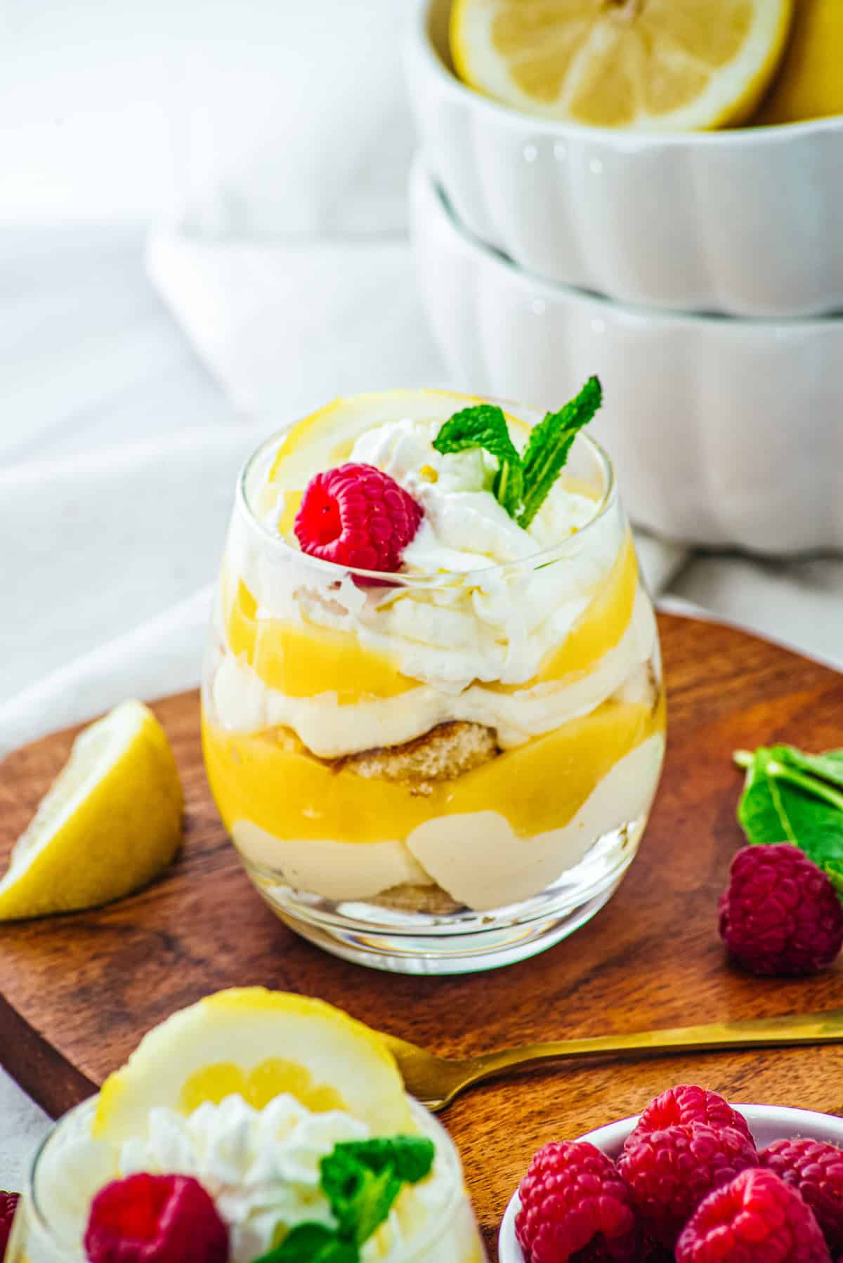 A glass dessert featuring Lemon Cheesecake Trifle Cups with luscious lemon curd, whipped cream, raspberries, and mint, presented on a wooden board alongside fresh lemons.