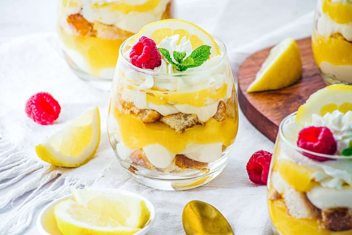 Glass of lemon cheesecake trifle cups dessert parfait topped with whipped cream, raspberry, mint, and lemon slice.
