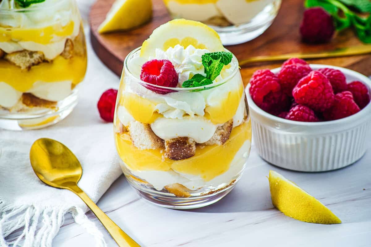 Glass of lemon dessert with whipped cream, mint, and raspberries beside a bowl of fresh raspberries—these Lemon Cheesecake Trifle Cups offer a delightful twist on classic flavors, beautifully layered and garnished for an elegant presentation.