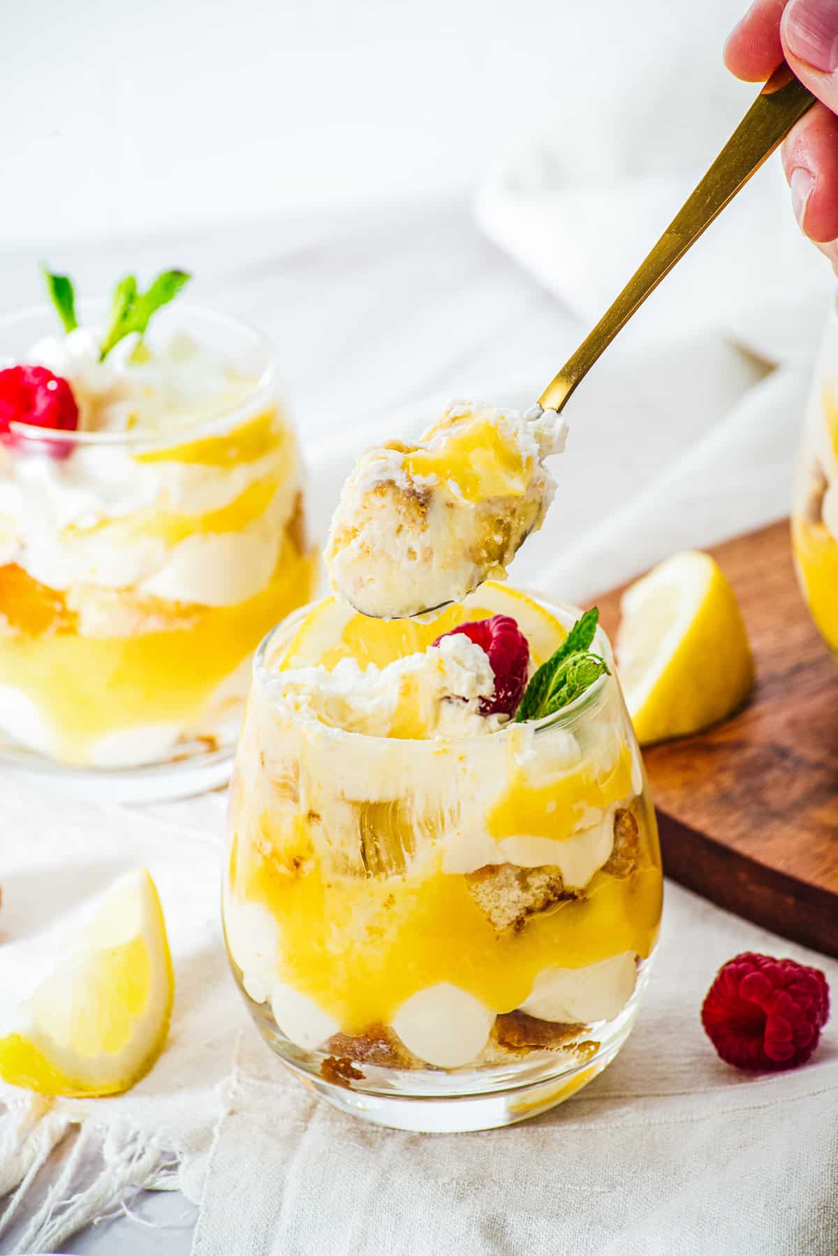 A glass dessert of Lemon Cheesecake Trifle Cups with lemon, cream, cake layers, topped with mint and raspberry. A spoon lifts a bite.