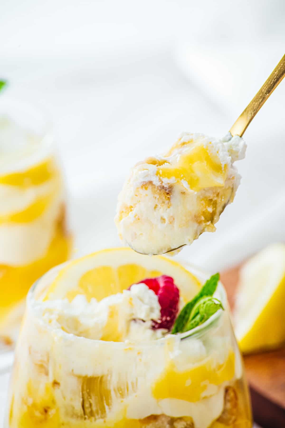 A spoonful of creamy lemon dessert with whipped topping, in front of a glass with layers of lemon and cream—perfect for enjoying these delightful Lemon Cheesecake Trifle Cups.