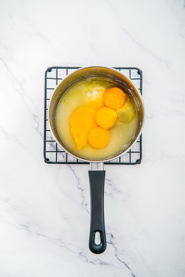 A saucepan with cracked eggs and sugar sits on a cooling rack over a white marble surface, ready to become the luscious base for indulgent Lemon Cheesecake Trifle Cups.