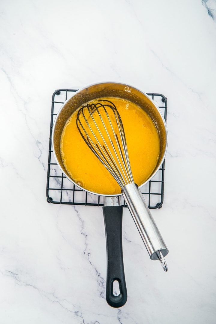 A saucepan with yellow sauce and a whisk on a cooling rack, set on a white marble surface, perfect for preparing the creamy base for Lemon Cheesecake Trifle Cups.