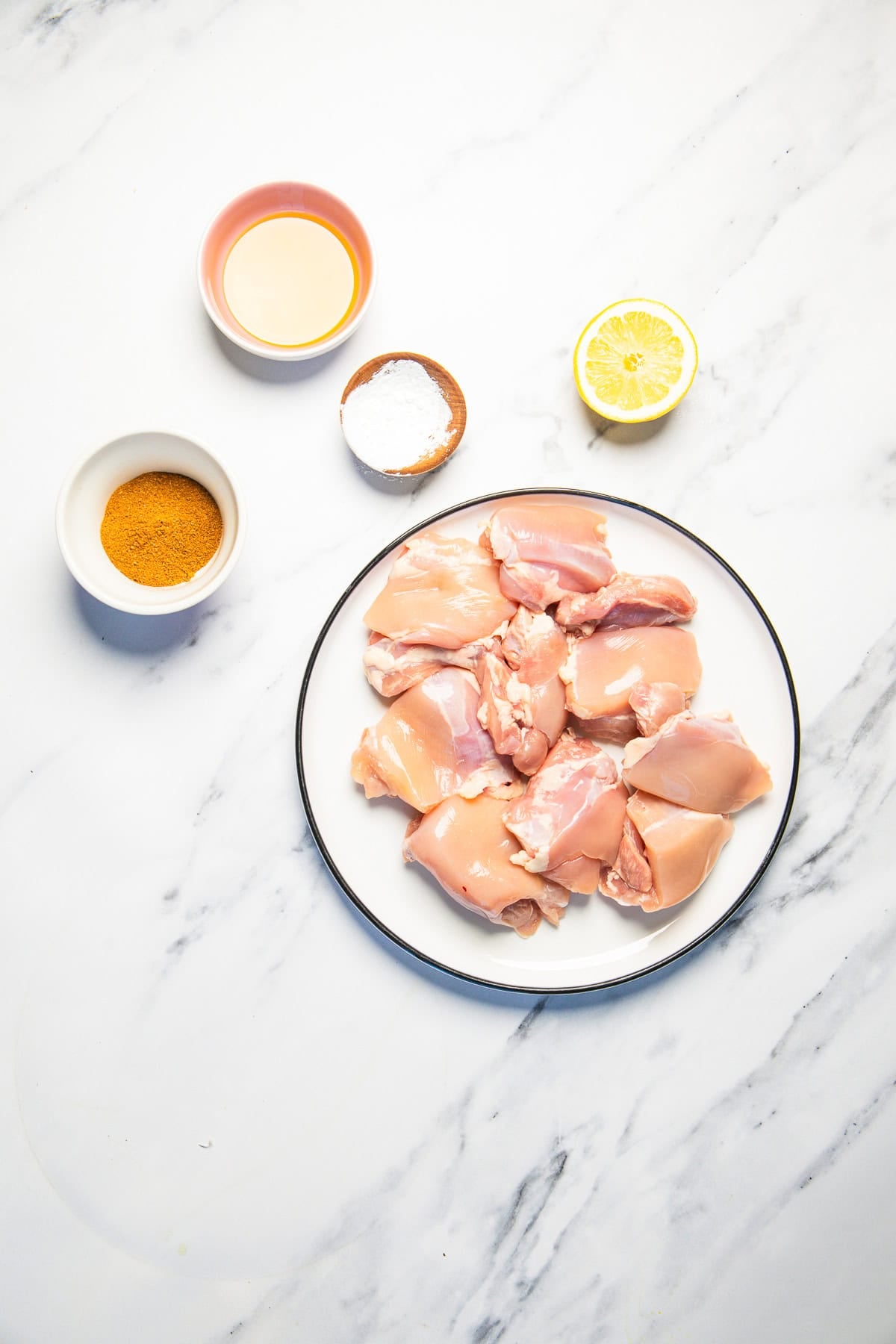 Raw chicken pieces on a plate with small bowls of spice, oil, flour, and a half lemon on a marble surface.