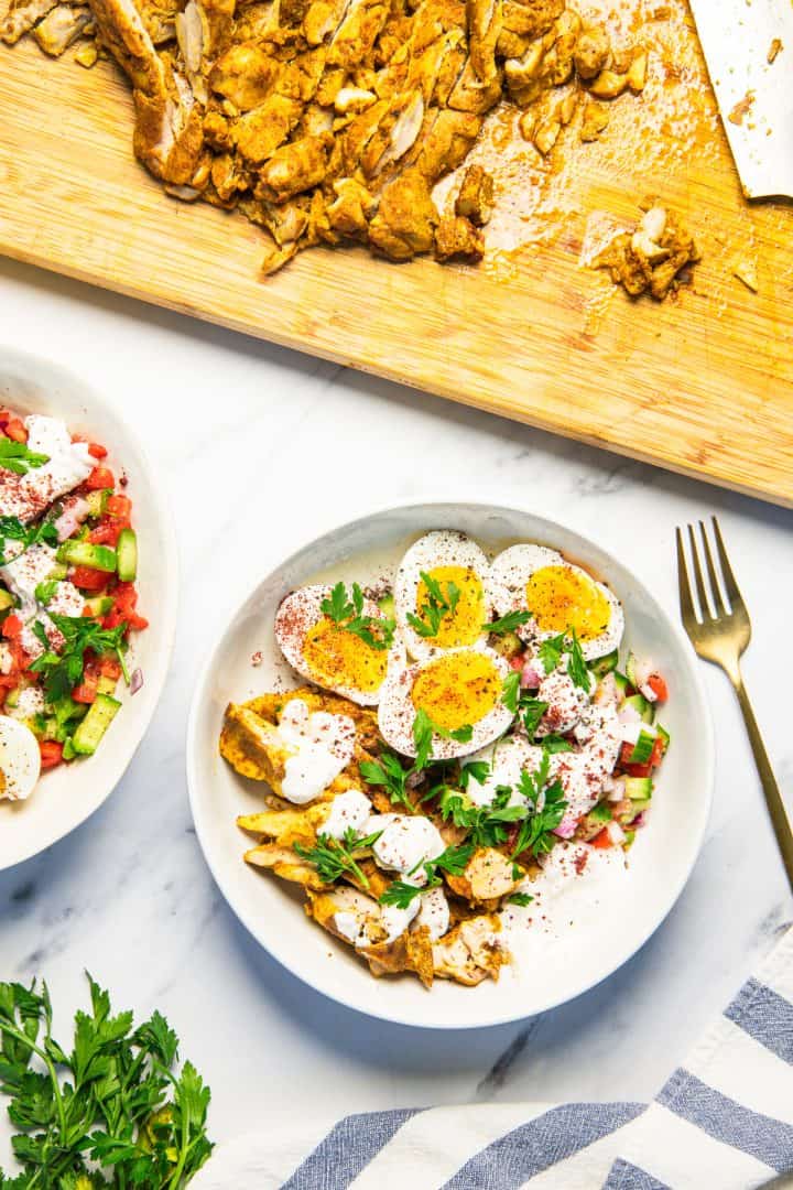 Bowl of Loaf Pan Chicken Shawarma salad with eggs, herbs, yogurt sauce, and a cutting board with sliced chicken.