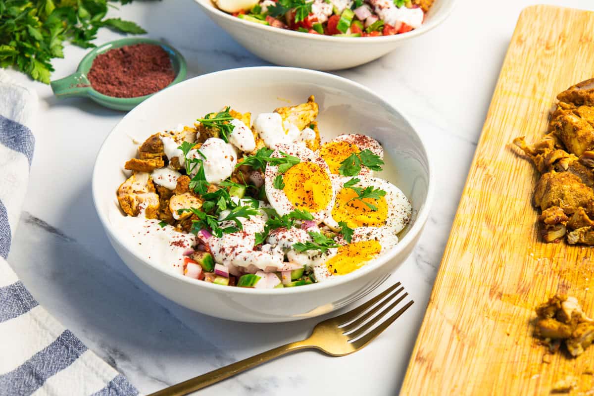 A bowl of salad with eggs, chicken, yogurt sauce, herbs, and spices, next to sliced chicken on a wooden board.