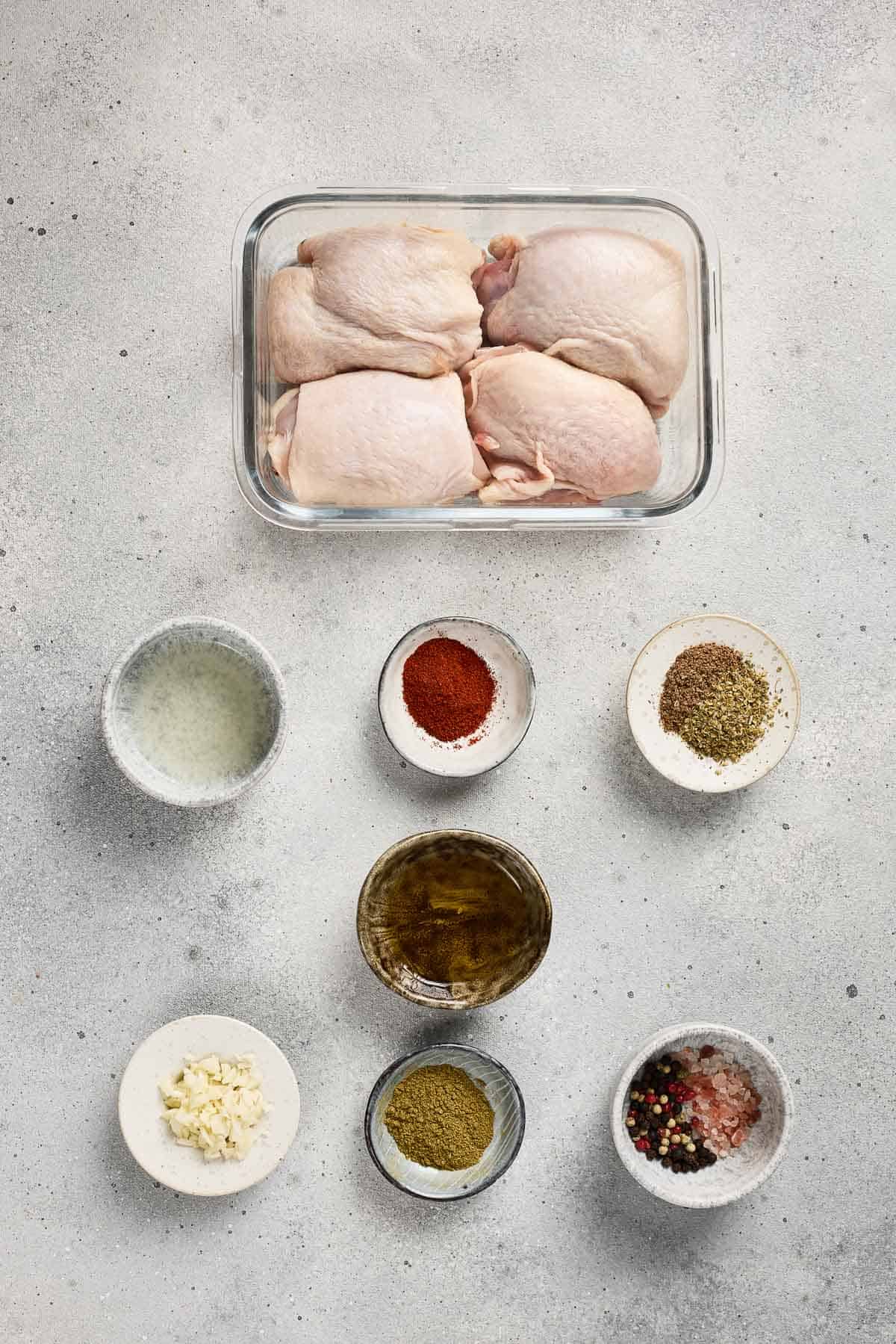 Raw chicken thighs in a glass dish with small bowls of spices, oil, minced garlic, and seasonings on a gray surface.