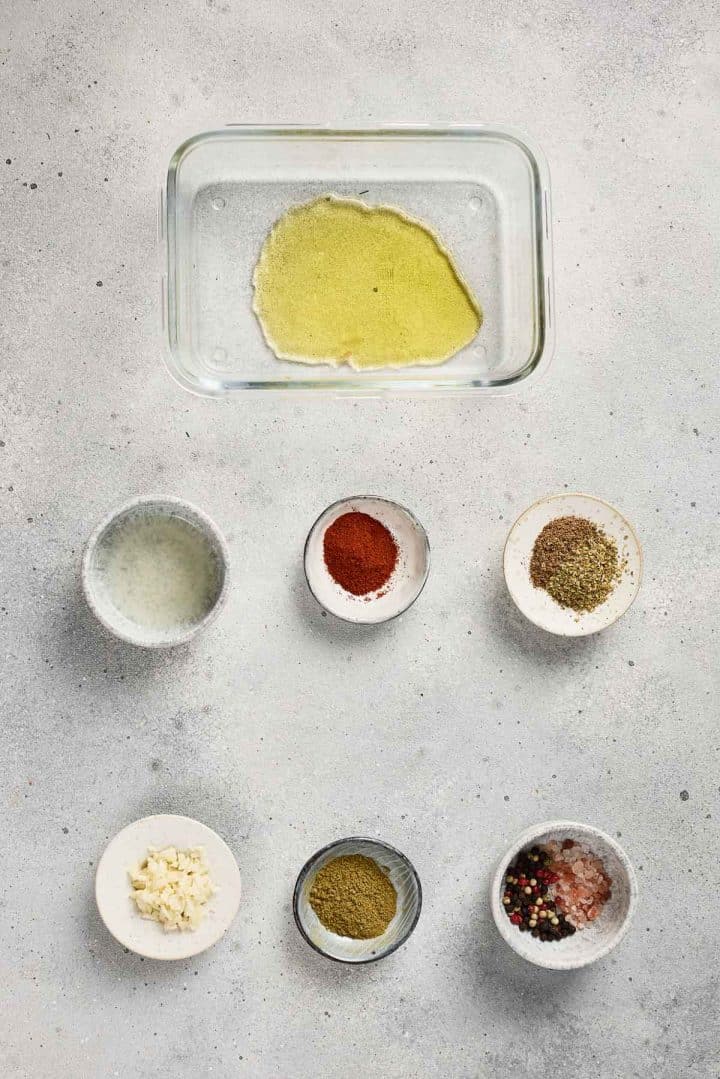 Glass dish with olive oil, surrounded by small bowls of spices, herbs, minced garlic, and lemon juice.