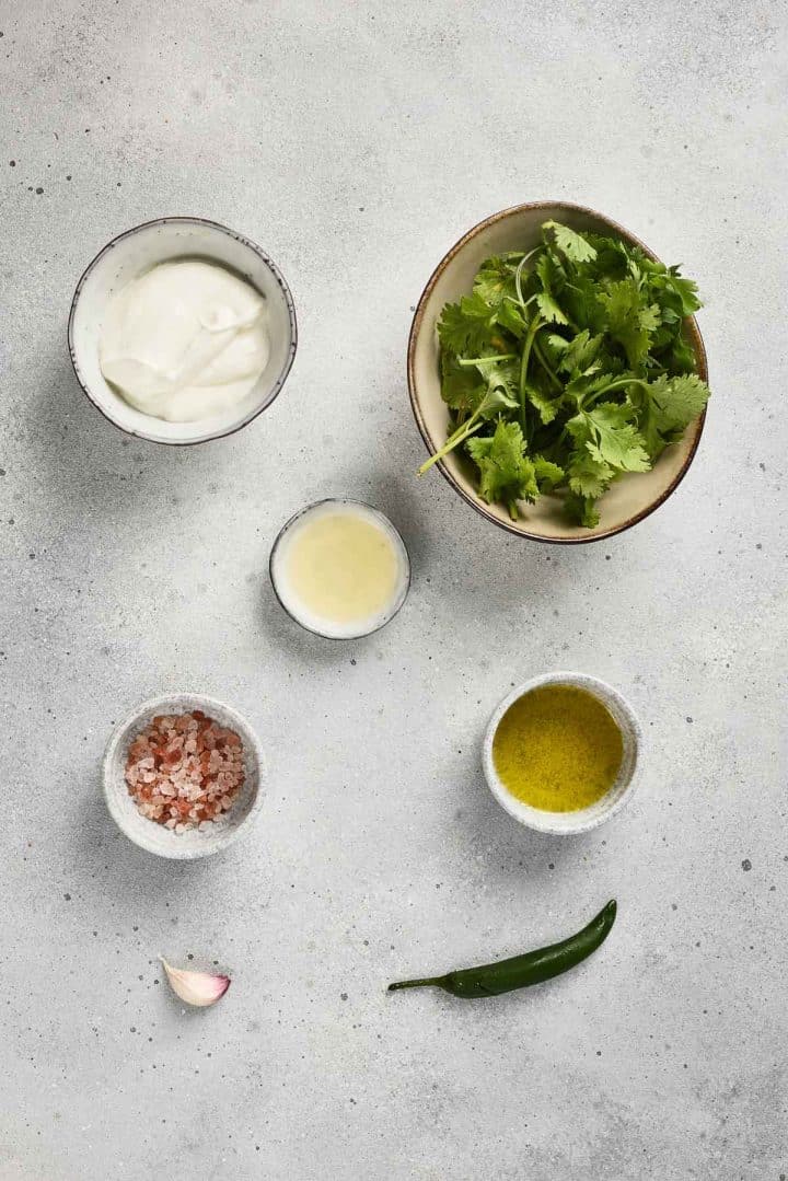 Five small bowls with yogurt, cilantro, lemon juice, salt, olive oil, plus a garlic clove and green chili on a gray surface.