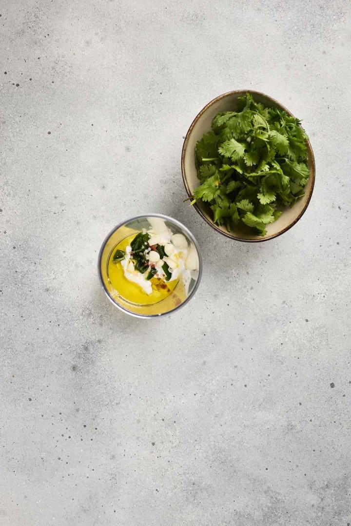 A bowl of fresh cilantro next to a glass of yellow sauce topped with herbs and sliced almonds on a gray surface.