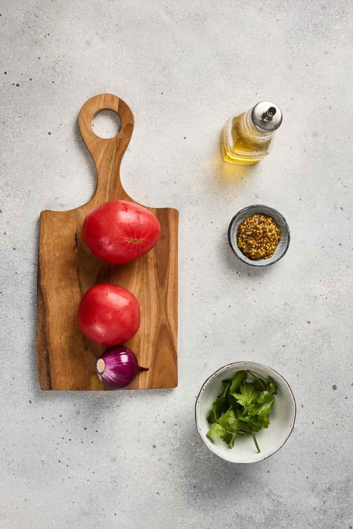 Two tomatoes and a red onion on a wooden board, with olive oil, spices, and cilantro on a light surface.