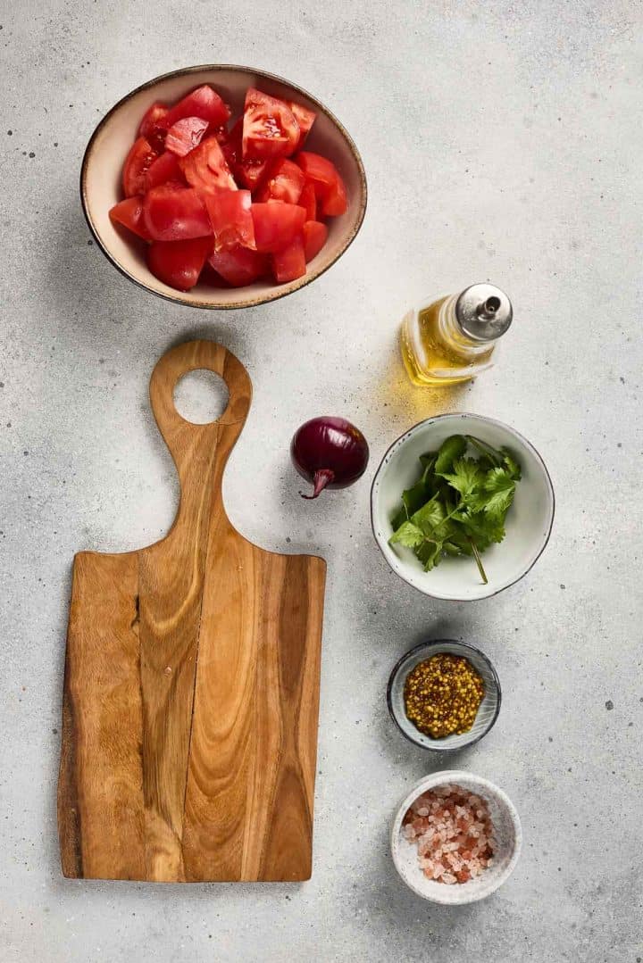 Chopped tomatoes, red onion, cilantro, oil, spices, and salt on a gray surface with a wooden cutting board.
