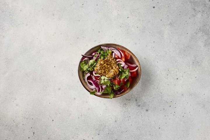 A bowl of salad with tomatoes, red onions, cilantro, and dressing on a light gray surface.