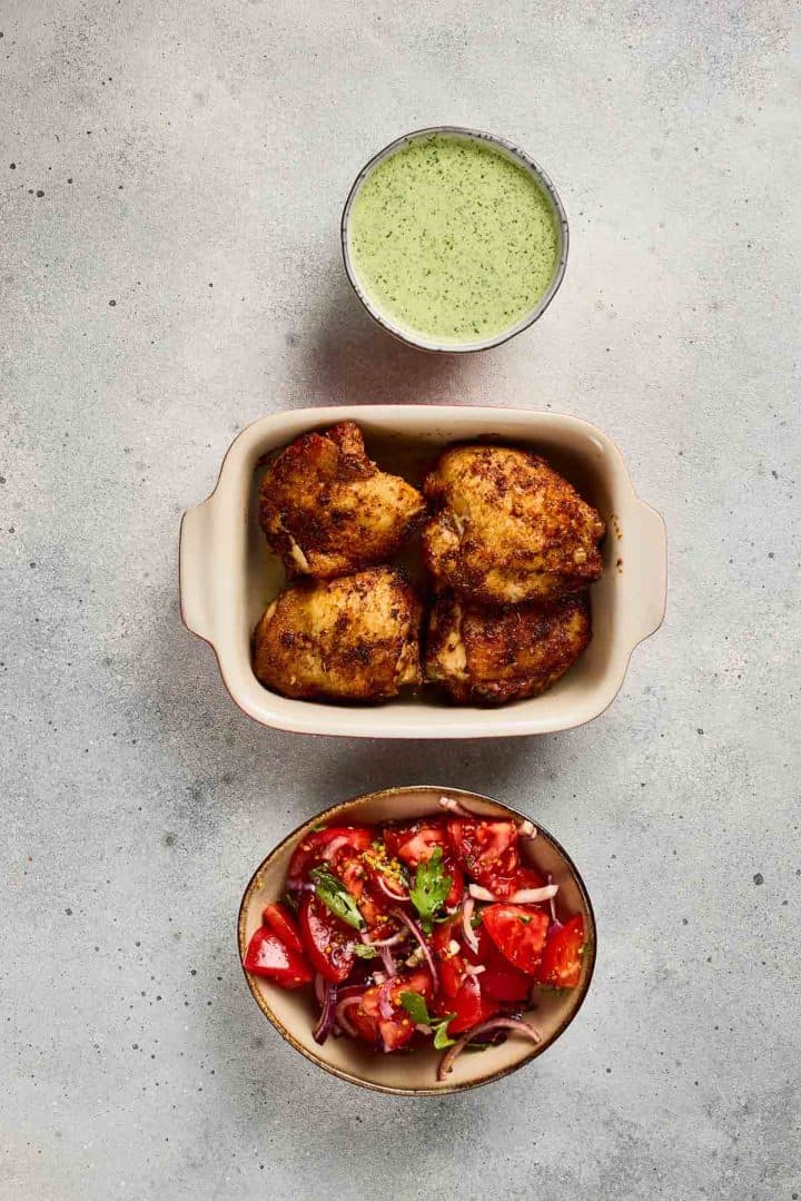 Three spiced chicken thighs, a bowl of tomato salad, and a cup of green sauce arranged on a light surface.