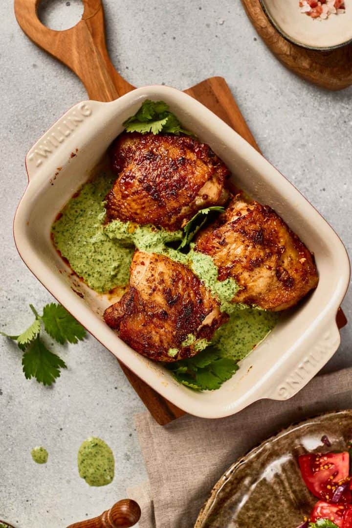 Three grilled chicken thighs sit on green sauce in a white baking dish, garnished with fresh cilantro.