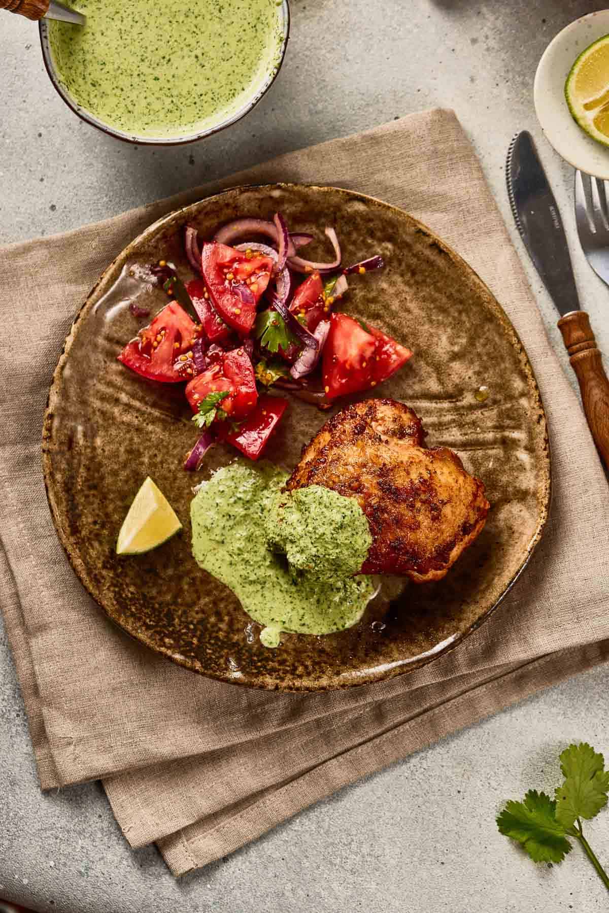 Brown plate featuring Peruvian Chicken with Green Sauce, served alongside a vibrant tomato salad and a lemon wedge on a tan napkin.