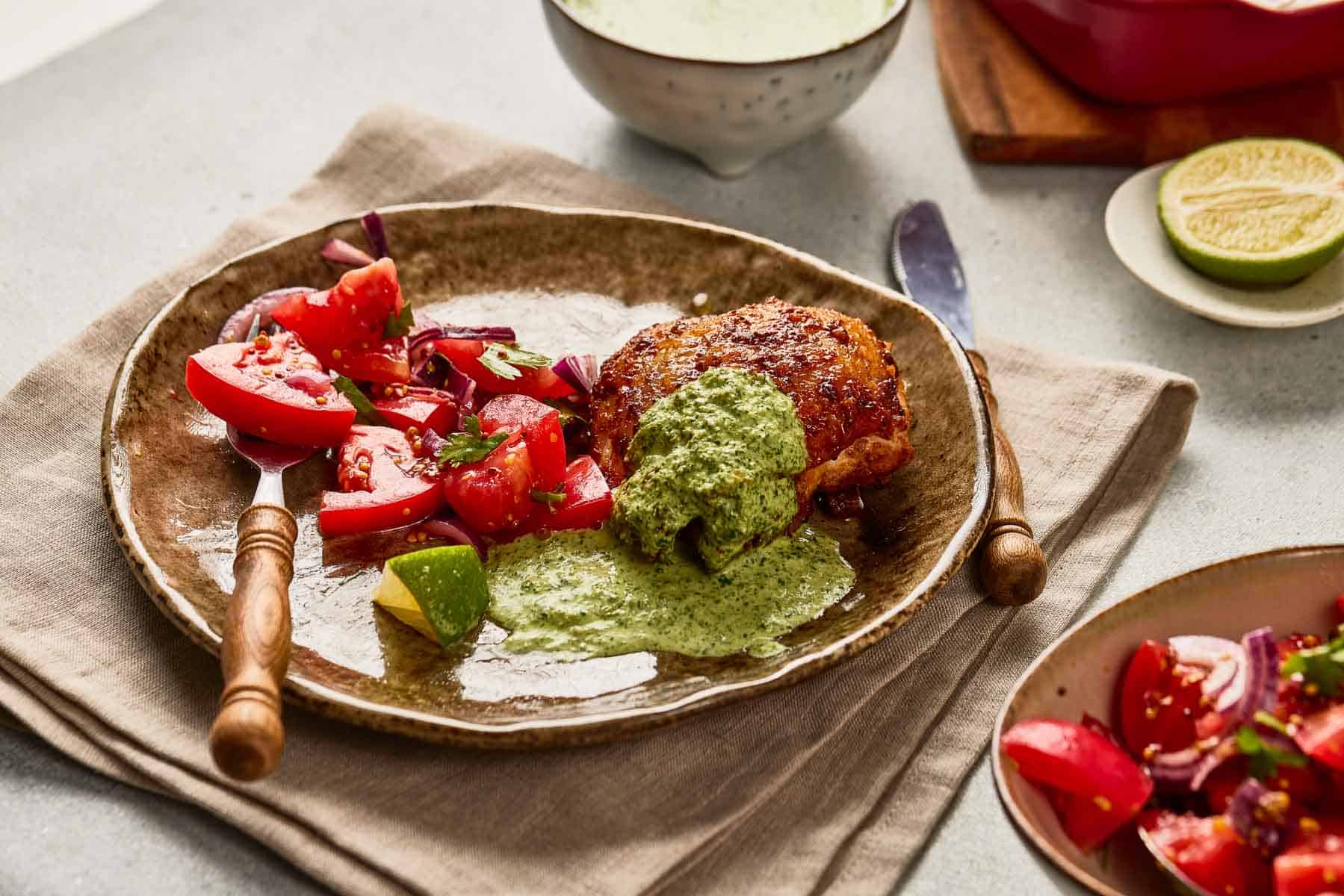 Brown plate with roasted chicken topped with green sauce, tomato salad, lime wedge, and a fork on a beige napkin.