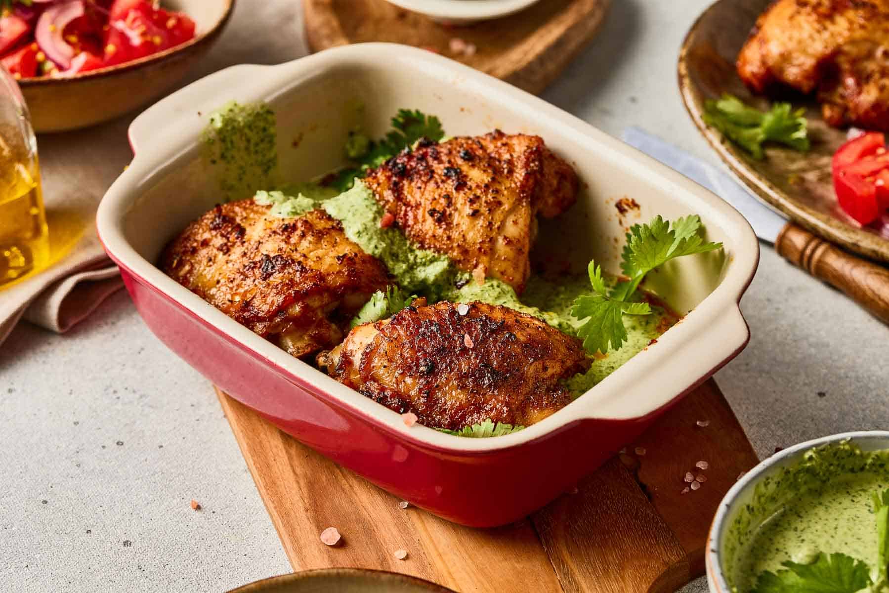 Peruvian Chicken with Green Sauce: Roasted chicken thighs in a red baking dish topped with vibrant green sauce and fresh cilantro on a wooden board.
