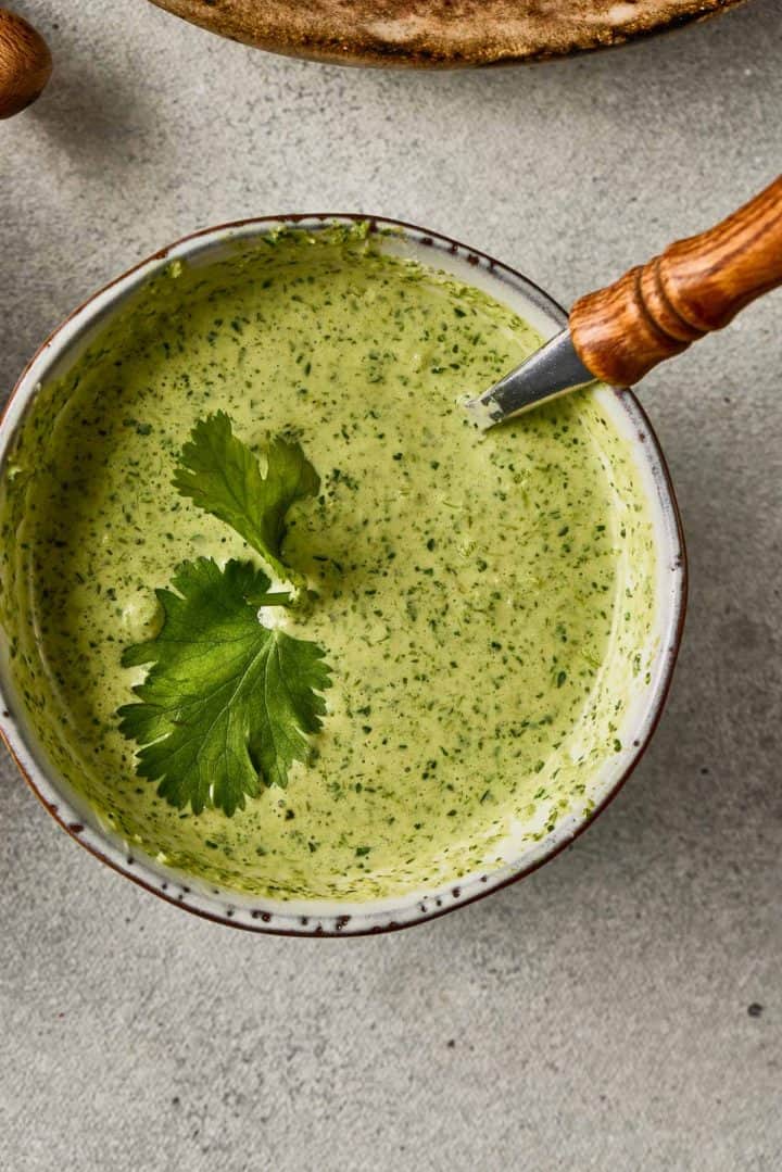 Bowl of creamy green cilantro sauce with a cilantro leaf garnish and a spoon on a gray surface.