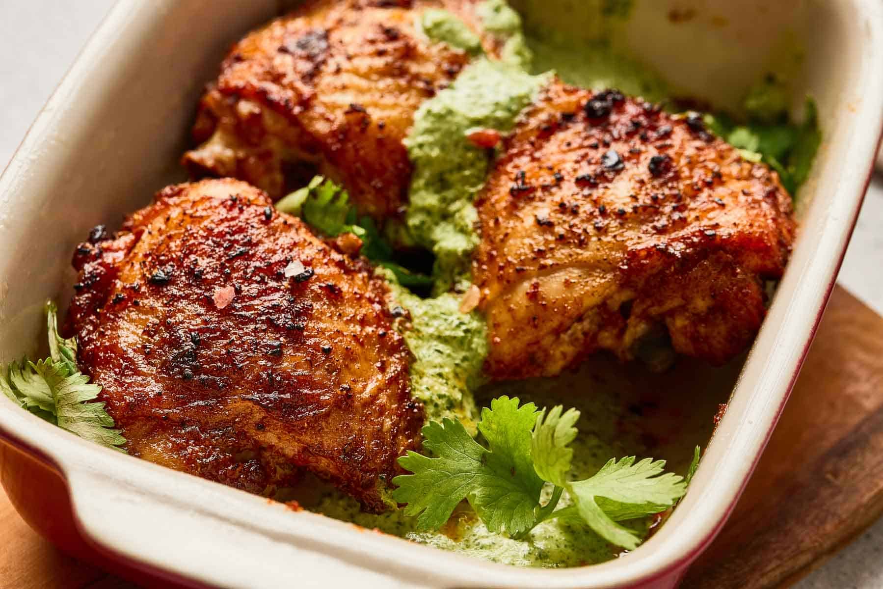 Three seasoned, roasted chicken thighs in a baking dish with green sauce and fresh cilantro garnish.