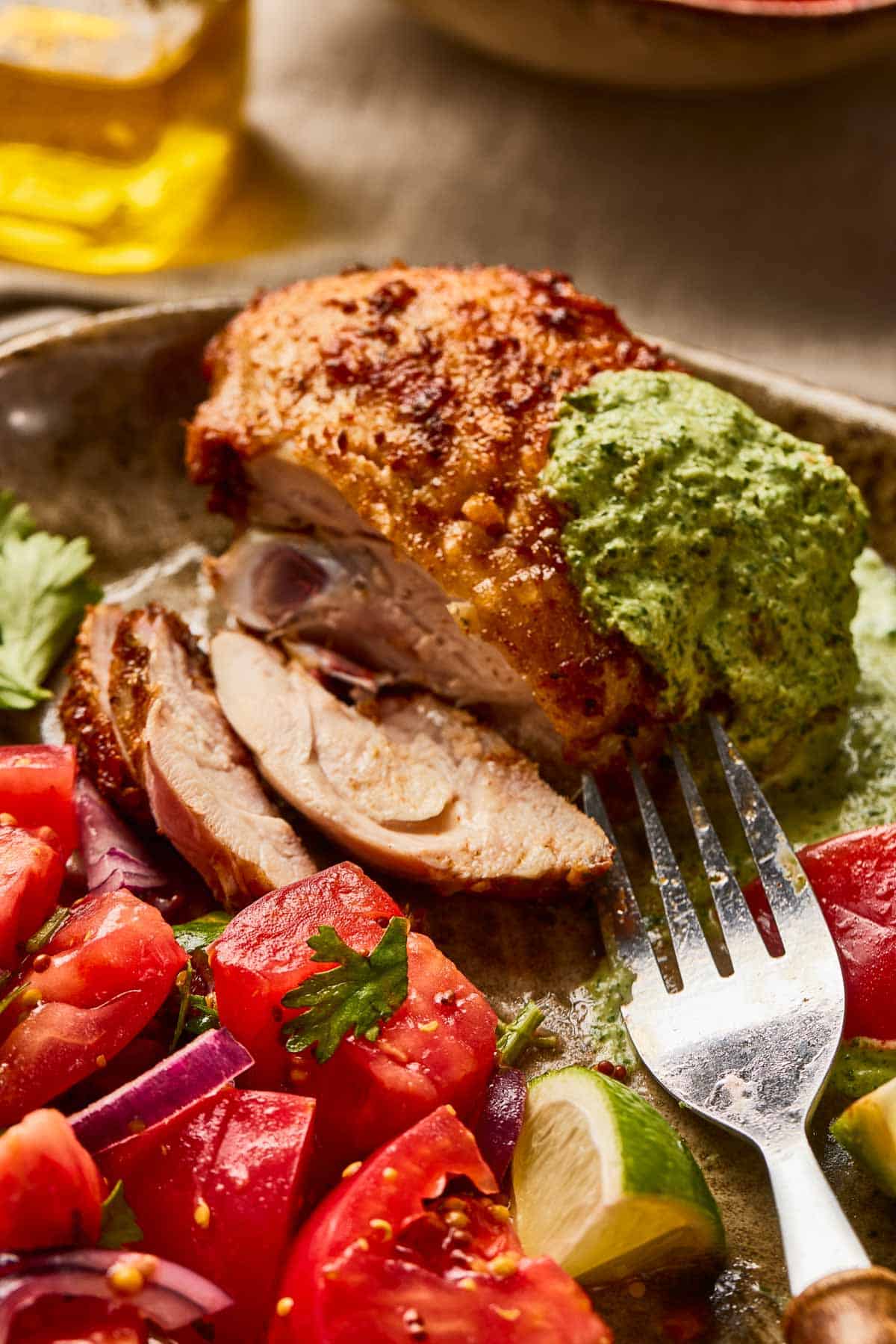 Close-up of seasoned chicken with green sauce, sliced tomatoes, red onions, and a fork on a plate.