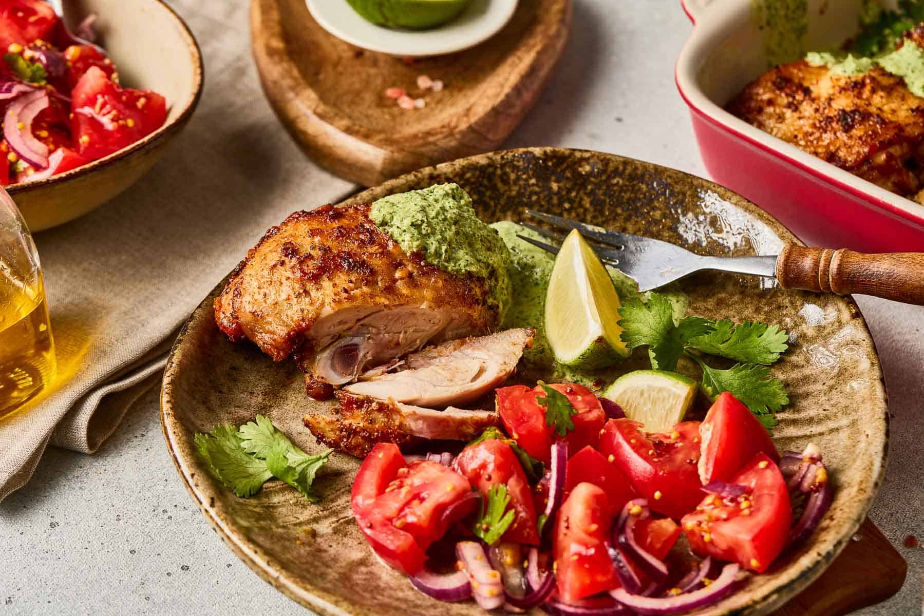 A plate of Peruvian Chicken with Green Sauce, served alongside a fresh tomato salad, cilantro, and lime wedges.