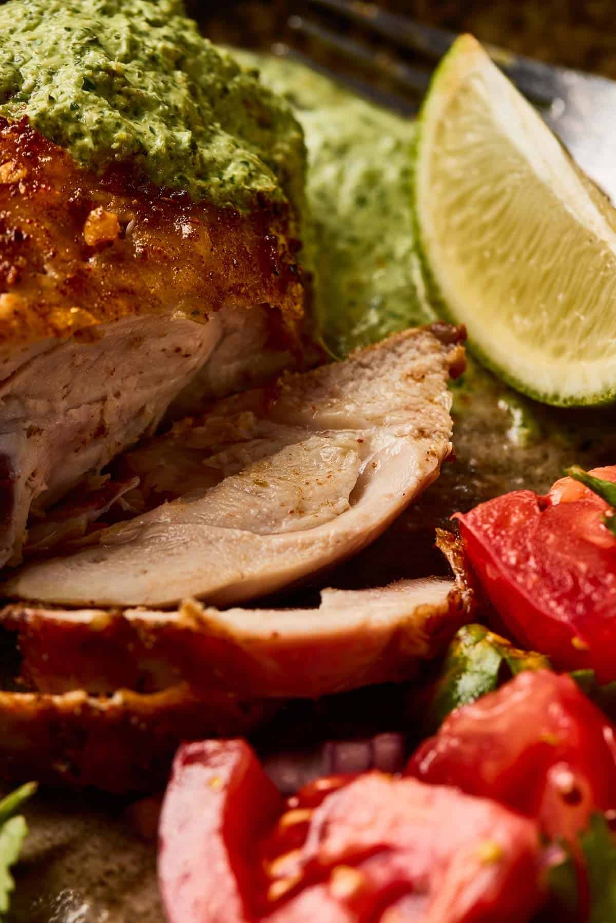 Sliced roast chicken with green sauce, lime wedge, and chopped tomatoes on a plate.