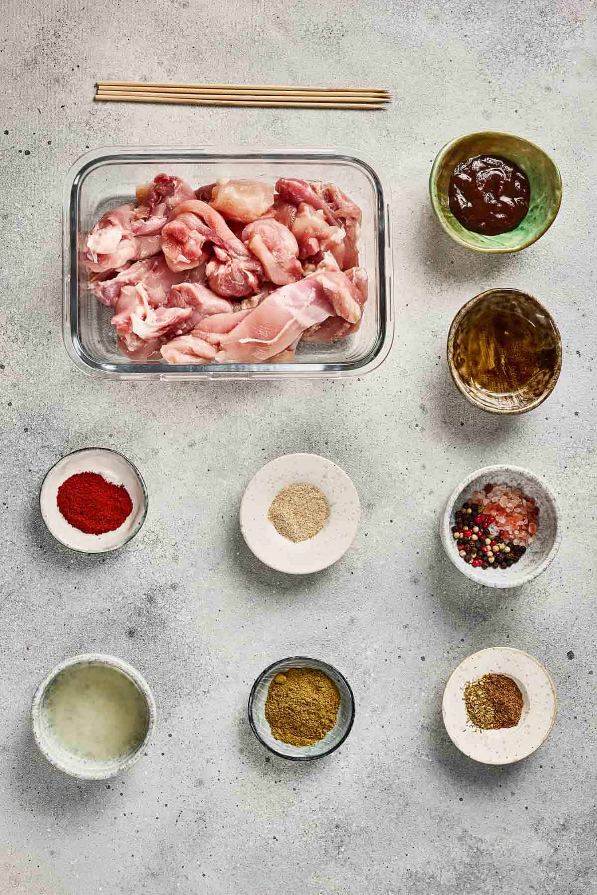 Raw chicken pieces in a glass container surrounded by small bowls of spices, sauces, and seasonings on a gray surface.