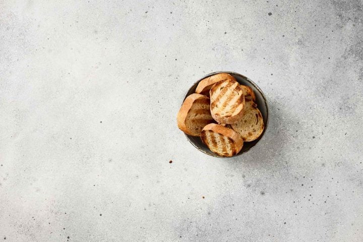 A small bowl filled with toasted bread slices on a light gray, textured surface.