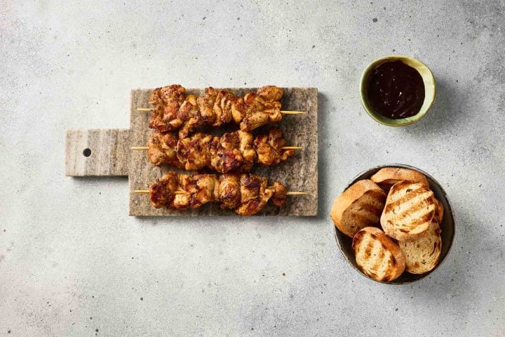 Two grilled meat skewers on a board, with a bowl of grilled bread slices and a small bowl of dark sauce nearby.