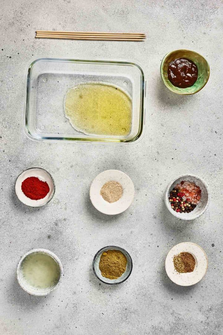 Glass dish with oil, seven small bowls of spices, sauce, and skewers arranged on a light gray surface.