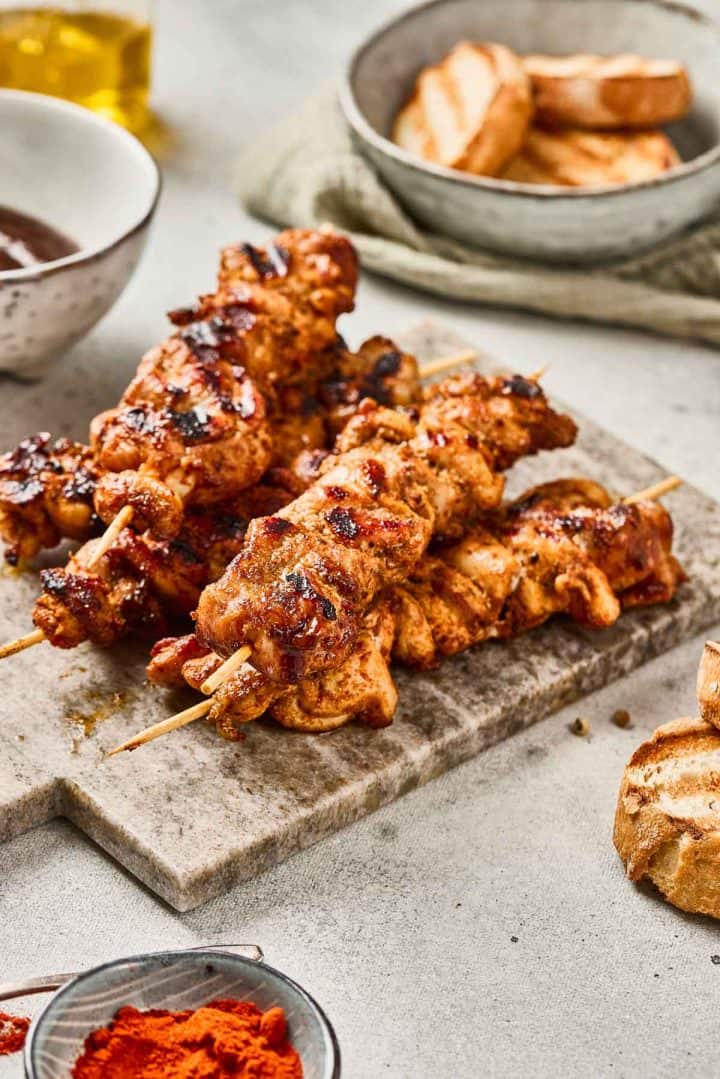 Grilled chicken skewers on a cutting board with grilled bread and bowls of sauce and spices nearby.