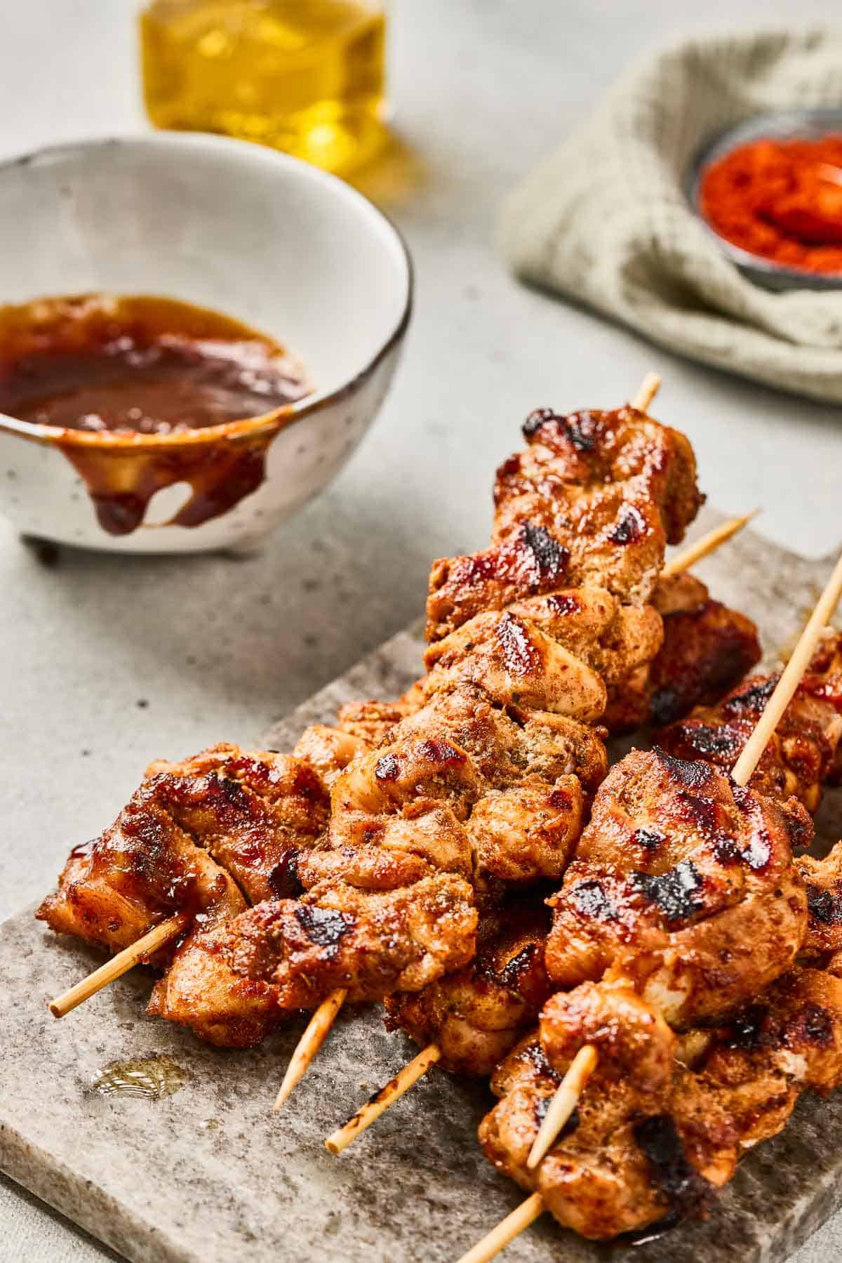 Grilled chicken skewers on a stone board with a bowl of sauce and spices in the background.