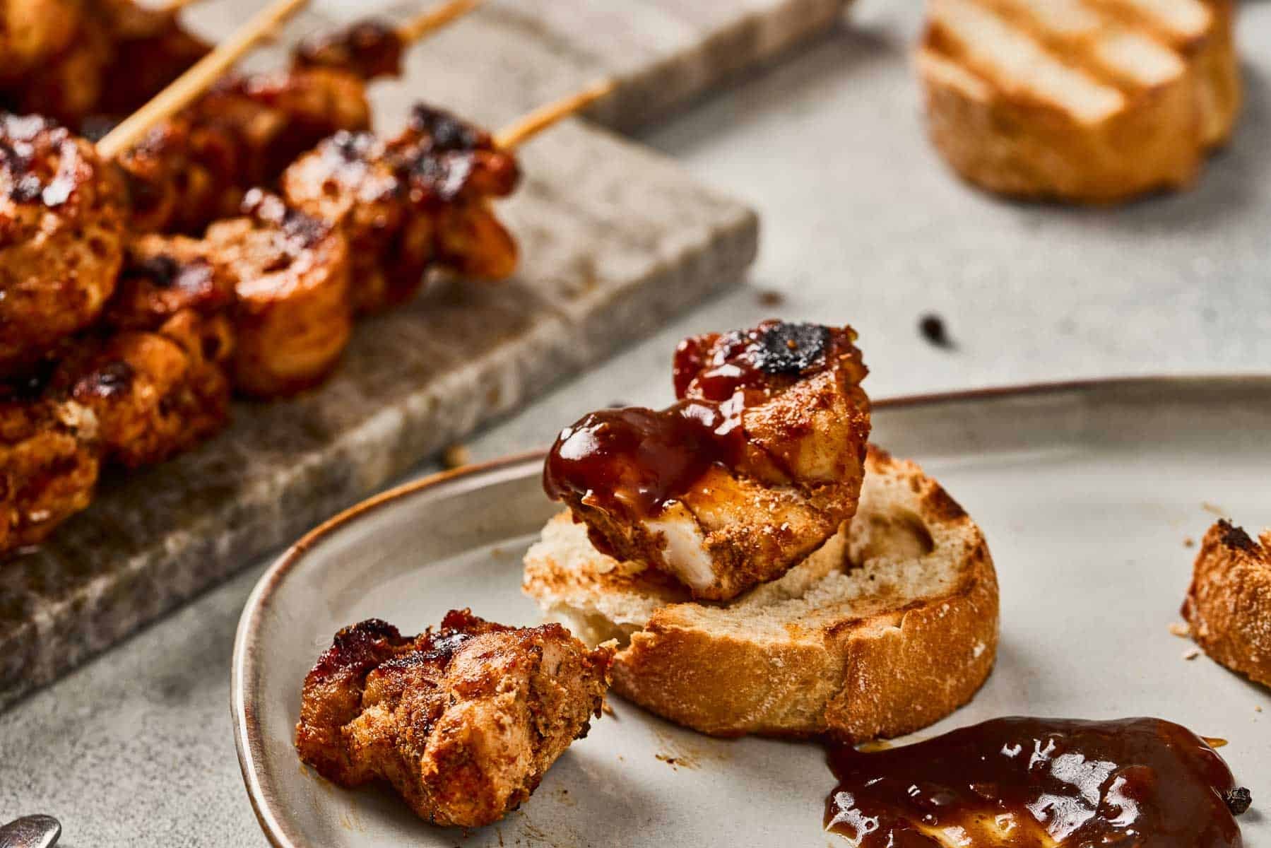 Grilled chicken skewers and sliced bread with barbecue sauce on a plate and marble board.