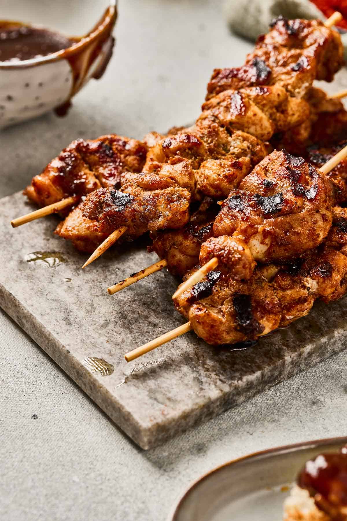 Grilled pinchos (Puerto Rican chicken skewers) served on a stone board, with barbecue sauce in a bowl in the background.