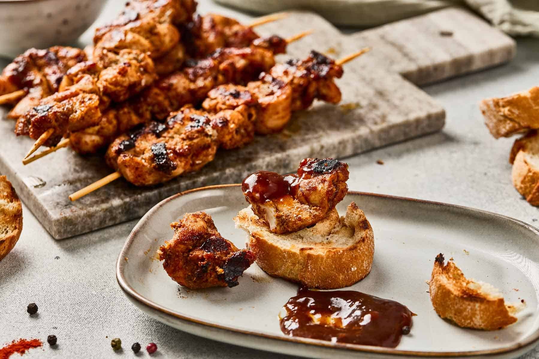 Grilled chicken skewers with bread slices and barbecue sauce on a plate and cutting board.