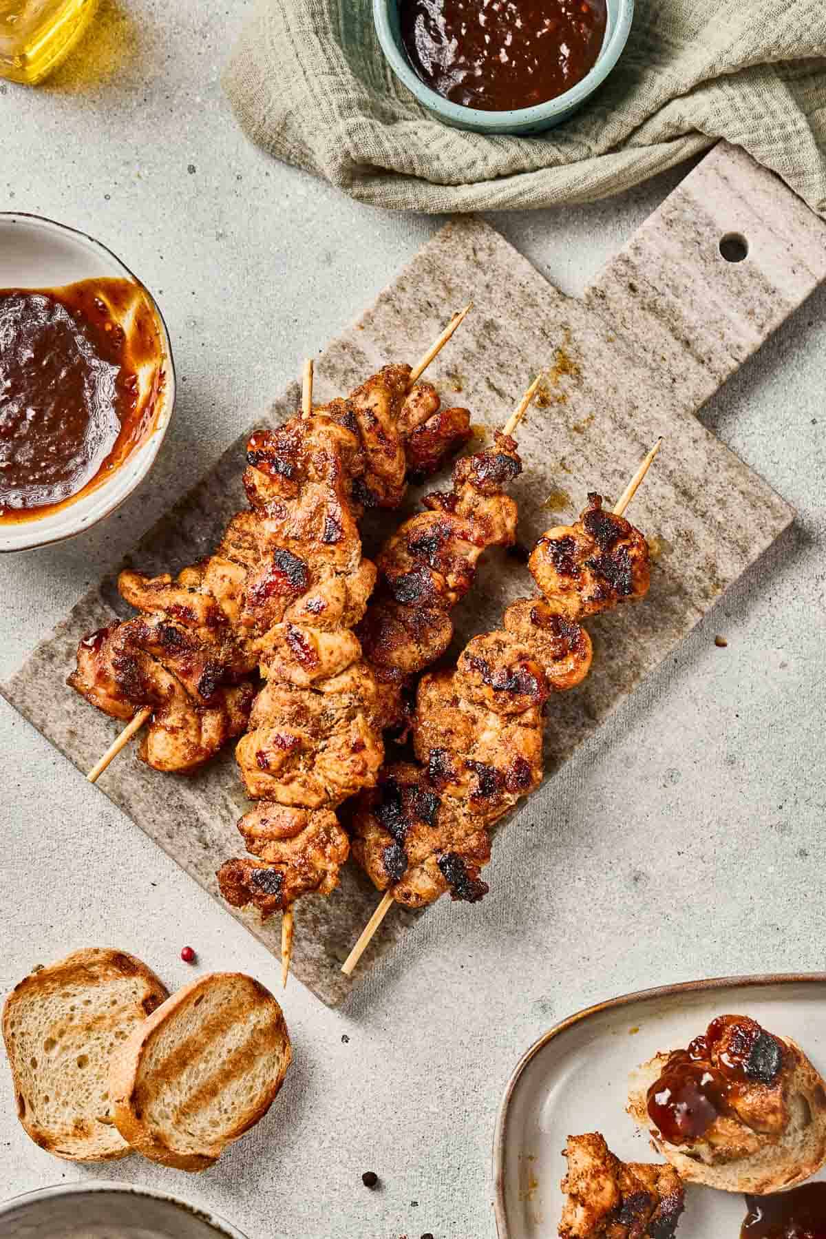 Grilled chicken skewers on a board with barbecue sauce, bread slices, and a napkin nearby.