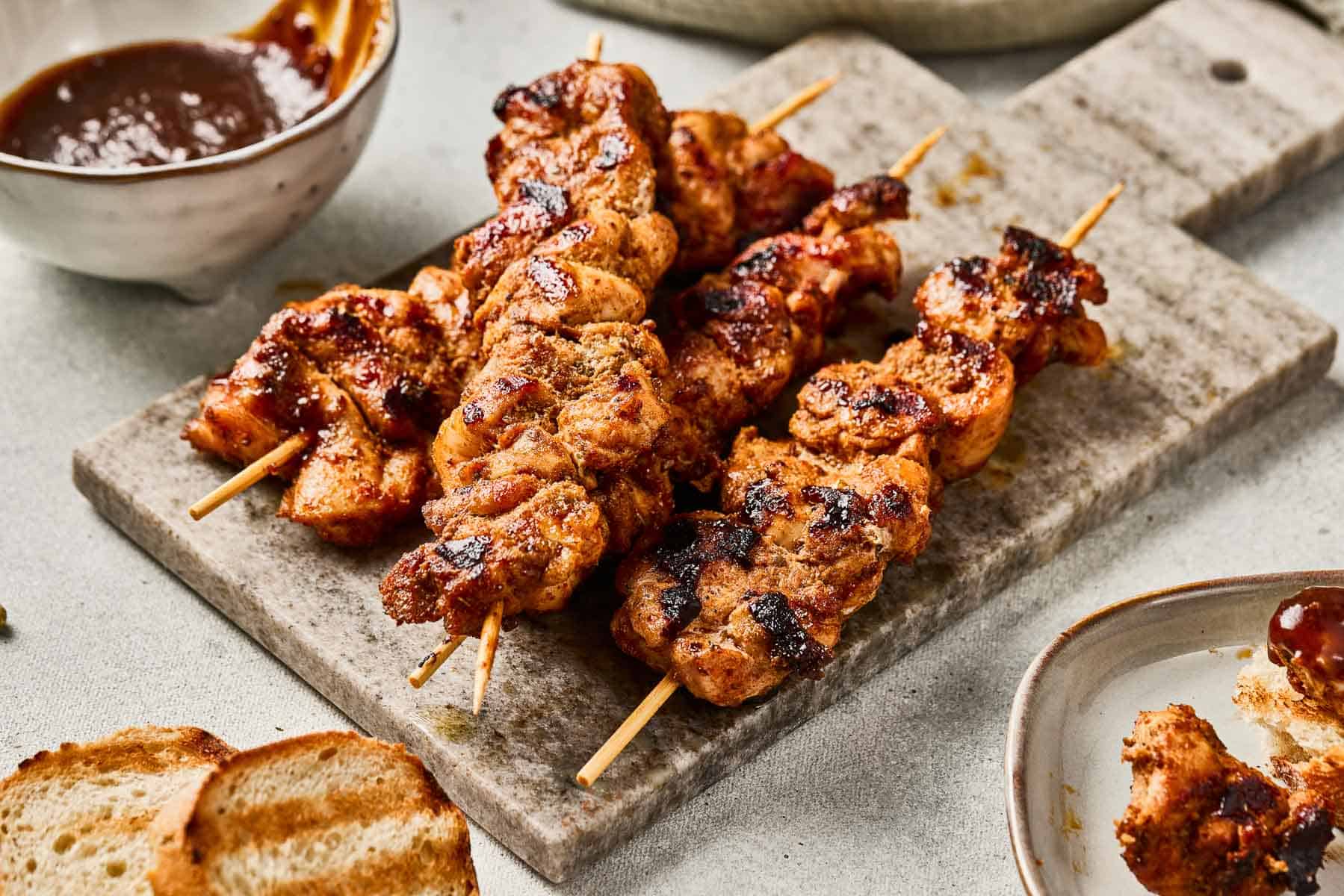Grilled chicken skewers on a board with barbecue sauce and toasted bread nearby.