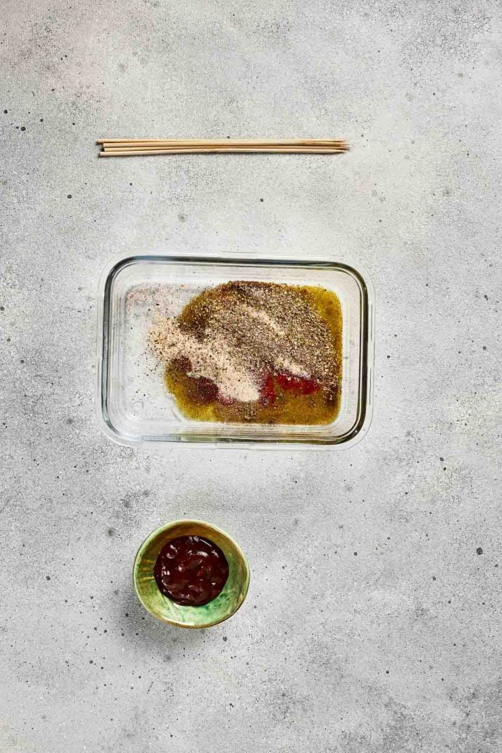 Glass dish with raw chicken and spices, a bowl of sauce, and chopsticks on a gray countertop.
