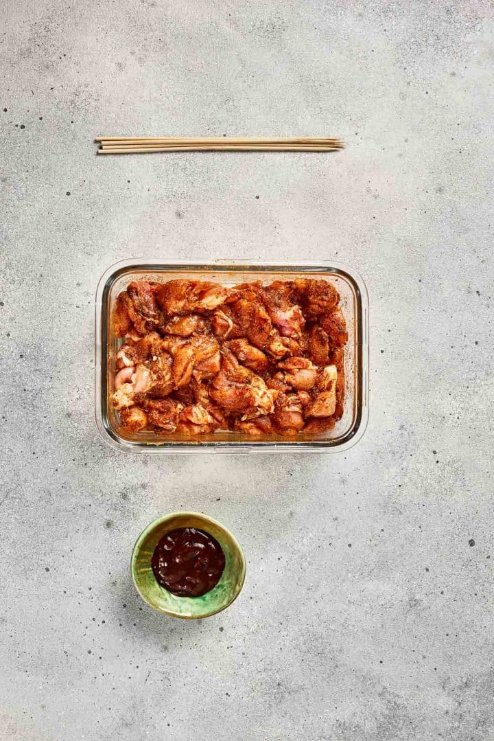 Glass container of marinated chicken pieces, bowl of sauce, and chopsticks on a gray surface.