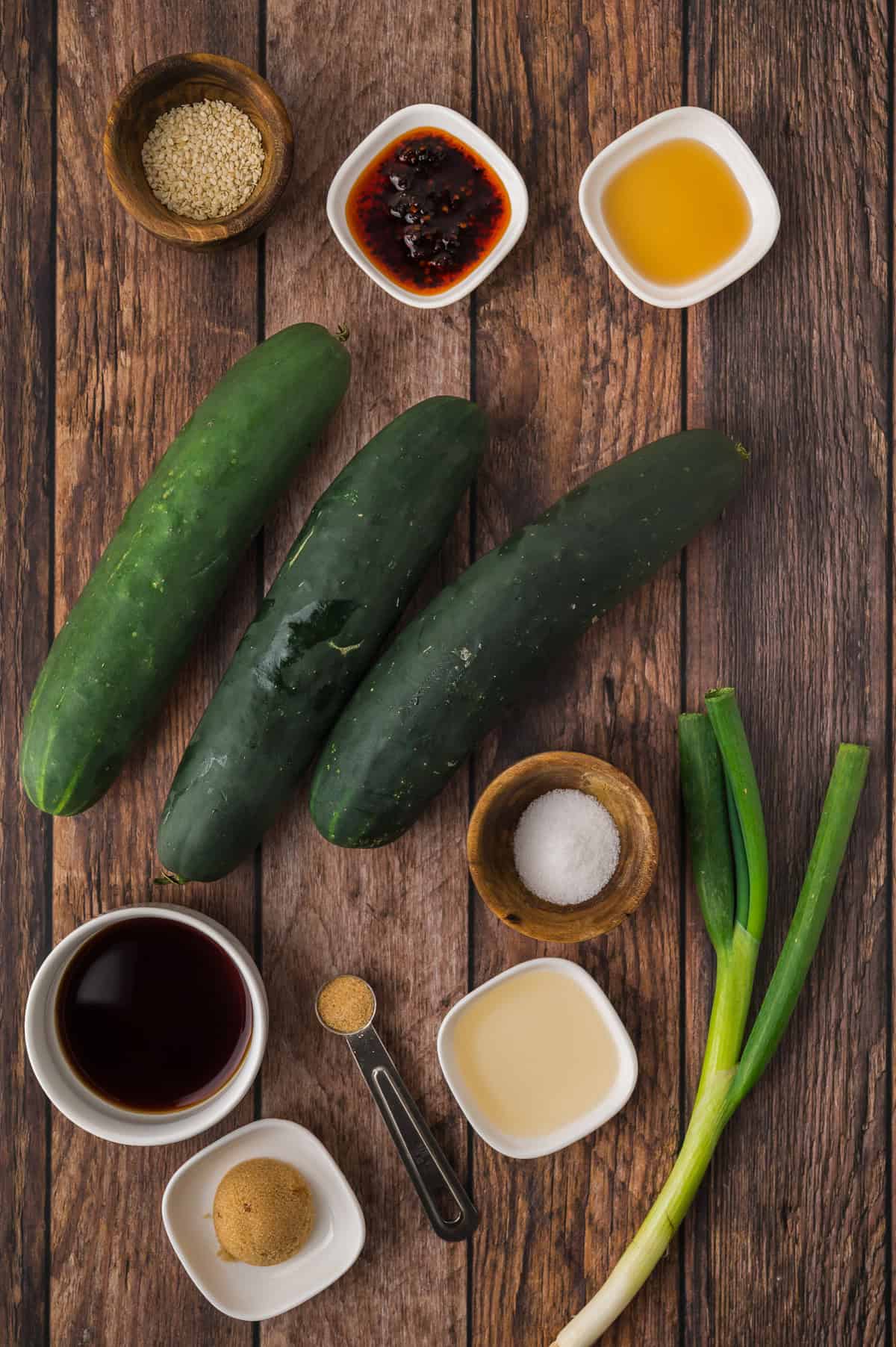 Flat lay of cucumbers, green onions, and small bowls with sauces, seasonings, and sugar on a wooden surface.