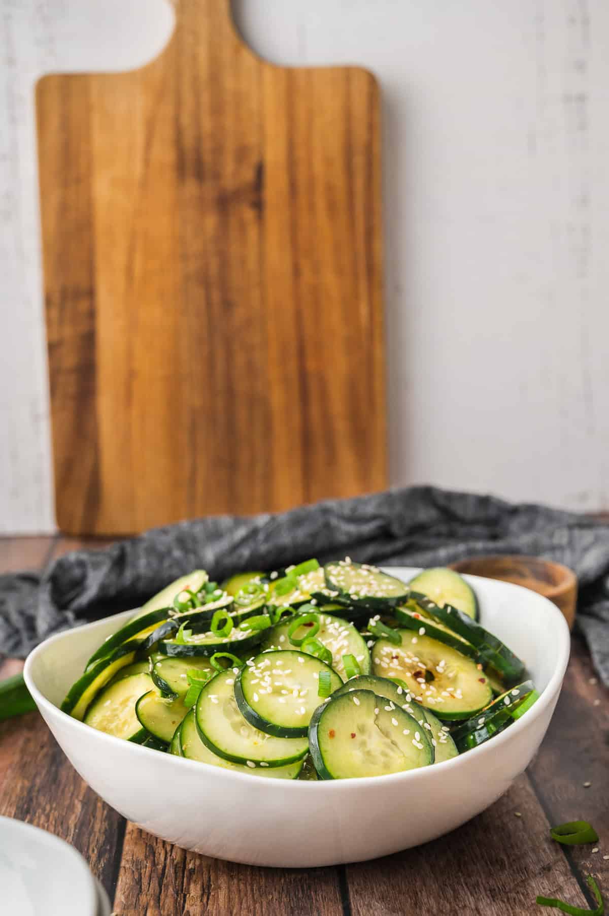 A white bowl of cucumber salad with sesame seeds, on a wooden table with a cutting board in the background.