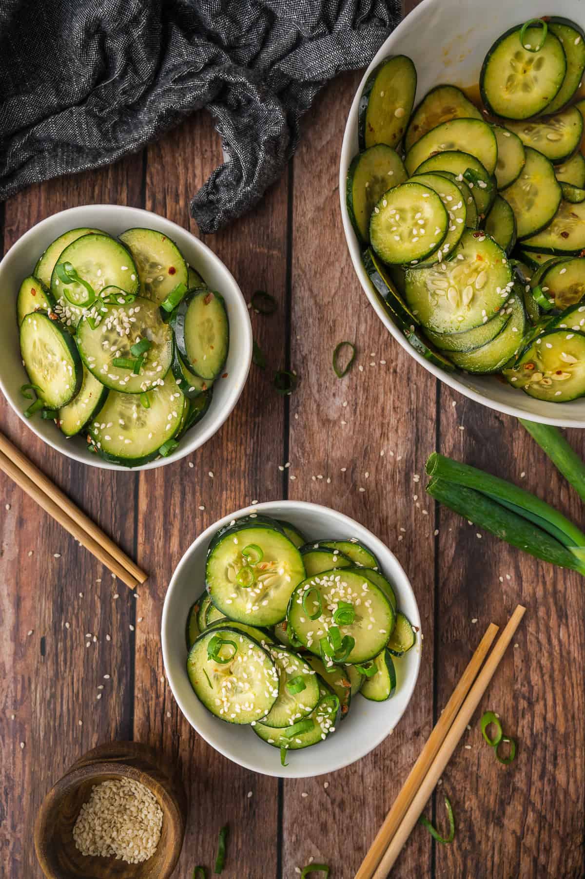 Bowls of sliced cucumber salad with sesame seeds and green onions on a wooden table, chopsticks beside them.
