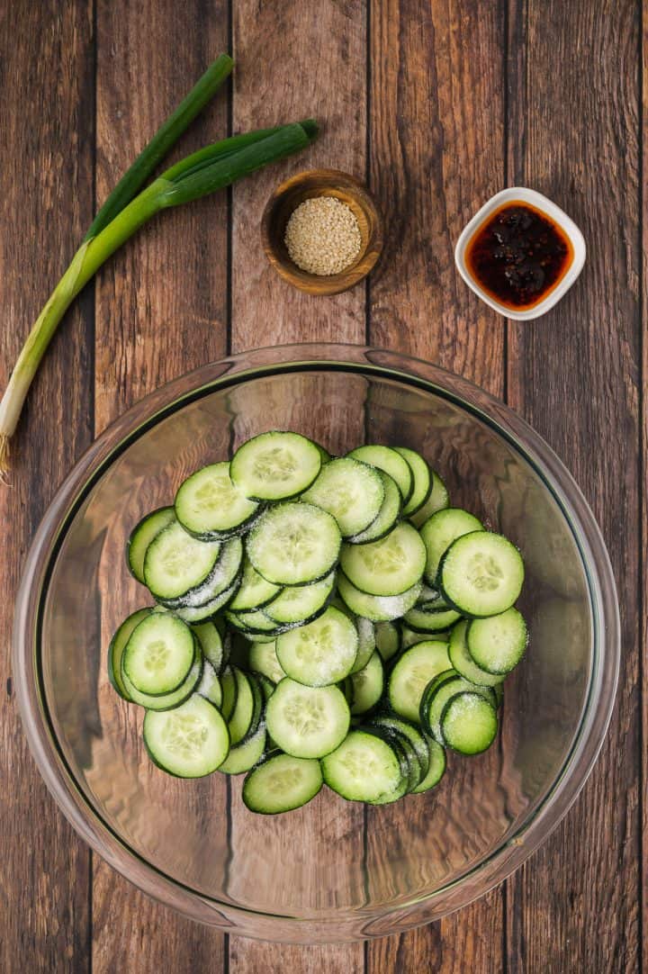 Sliced cucumbers in a glass bowl with salt, next to scallions, sesame seeds, and chili oil on a wooden table.