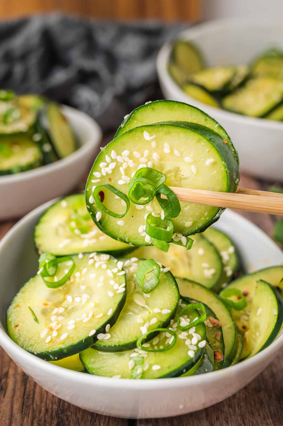 Chopsticks holding cucumber slices over a bowl of cucumber salad with sesame seeds and green onions.