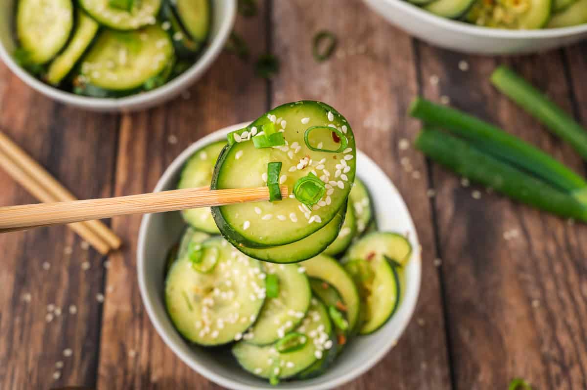 Chopsticks holding cucumber slices with sesame seeds and green onions over a bowl on a wooden table.