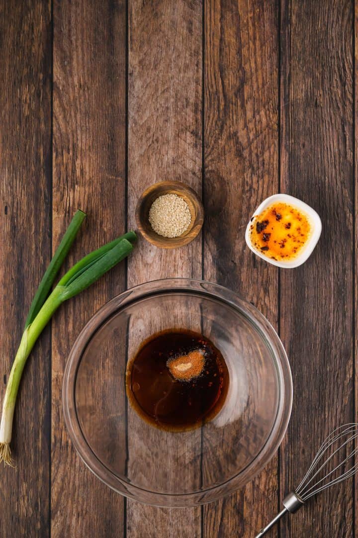 Glass bowl with sauce, sesame seeds, chili oil, scallion, and whisk on a wooden table, top-down view.